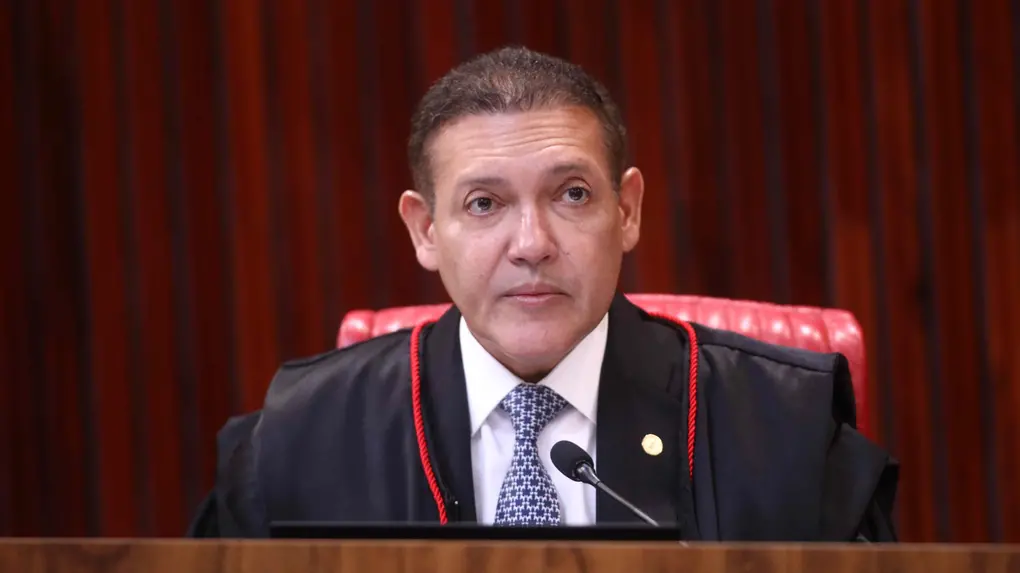 Homem de meia-idade, com cabelos curtos e escuros, sentado atrás de uma bancada de madeira, usando toga preta com detalhes em vermelho sobre terno, camisa branca e gravata estampada. Trata-se de Nunes Marques. Ele está em frente a um microfone, com expressão séria, e ao fundo há uma parede com painéis de madeira em tom avermelhado e uma cadeira estofada vermelha.