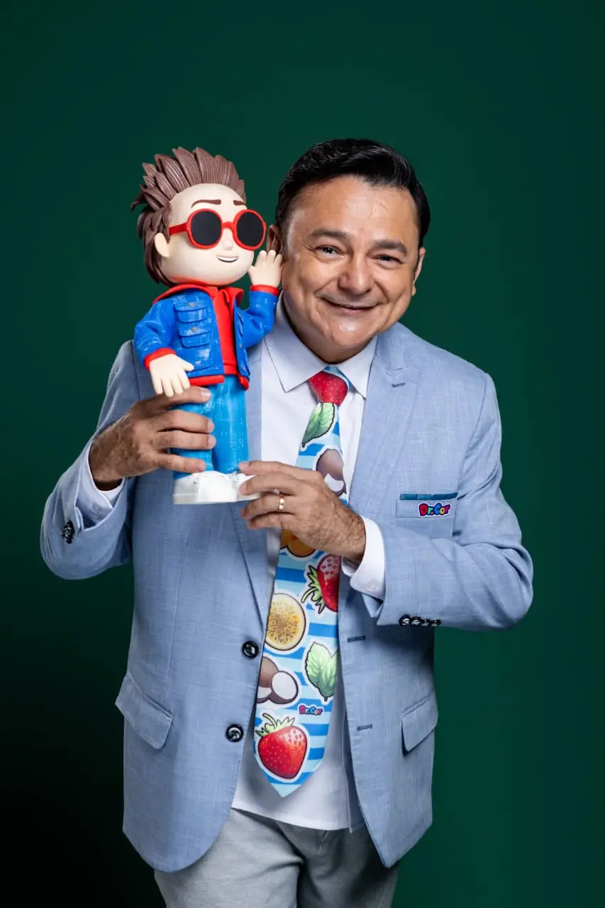 Homem de terno azul claro e gravata colorida sorri enquanto segura um boneco estilizado de óculos escuros e jaqueta azul, sobre fundo verde.