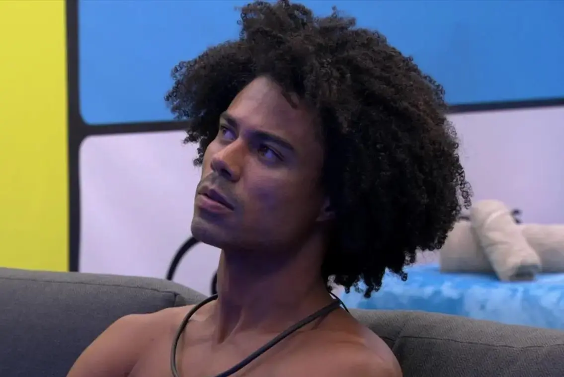 Uma pessoa sentada em um sofá de cor escura, em um ambiente interno iluminado. A pessoa possui cabelo volumoso e cacheado. Ao fundo, há uma parede azul e branca com formas geométricas, além de uma cama com cobertas e toalhas claras. A cena parece capturada em um espaço semelhante a uma sala ou quarto de programa de TV.