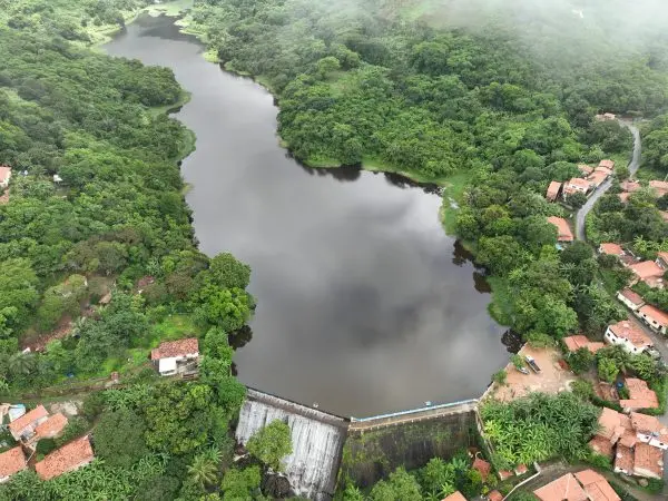 Vista aérea de uma represa cercada por densa floresta, com uma barragem visível e um pequeno vilarejo à margem na cidade de Baturité, no Ceará.