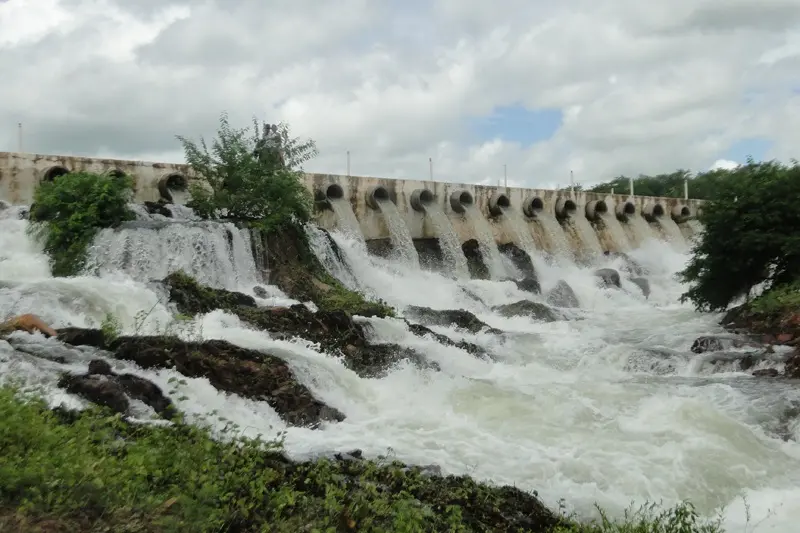 Vista da barragem Muquém liberando grande volume de água através de tubulações, formando corredeiras e cachoeiras espumantes sobre um leito rochoso, com vegetação verde ao redor e um céu nublado.