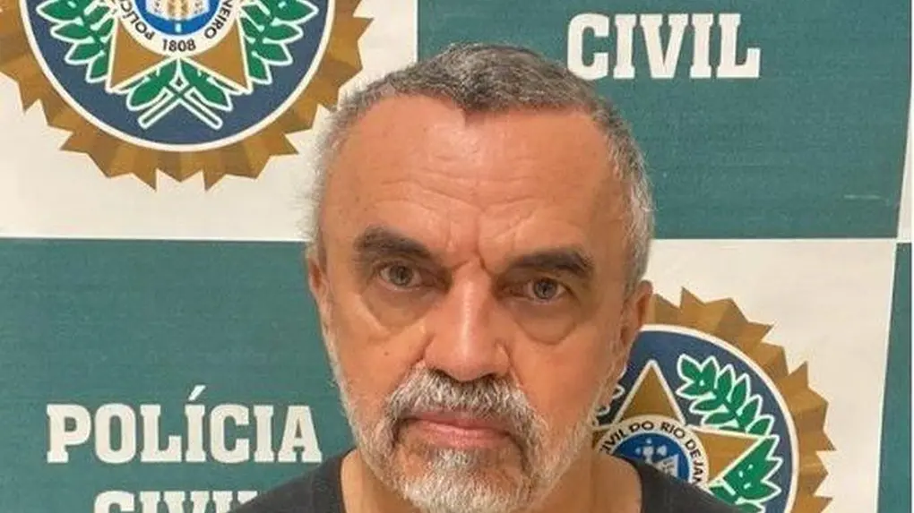 Ator José Dumont em retrato da prisão, olhando seriamente para a câmera em um plano médio. Ele está posicionado à frente de um painel institucional verde e branco da Polícia Civil do Rio de Janeiro.
