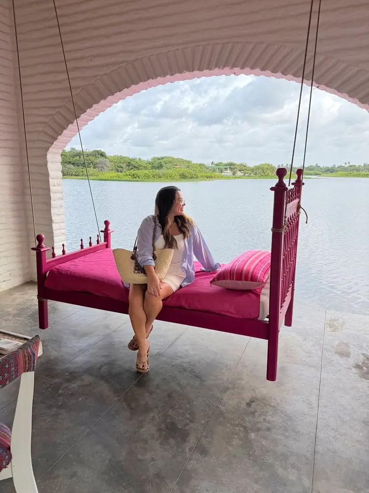 Na imagem, uma mulher jovem de cabelos castanhos longos está sentada em um leito de balanço suspenso de cor rosa vibrante (magenta). Ela está de perfil, olhando para a direita em direção a uma bela paisagem natural. Ela veste um vestido curto branco, uma camisa de botões azul clara aberta por cima e sandálias de salto. Ao seu lado, há uma bolsa de palha e um travesseiro listrado em tons de rosa.