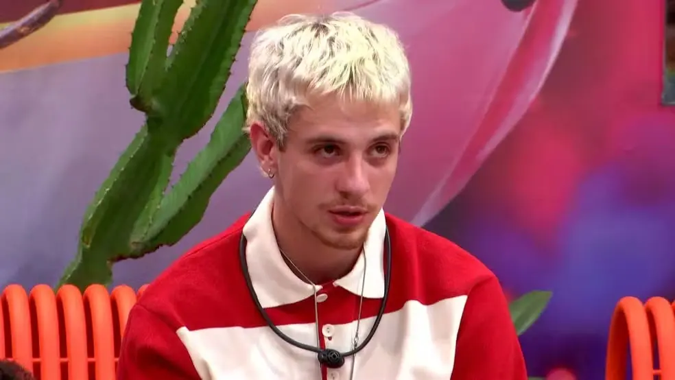 O influenciador Juliano Floss, com cabelos curtos e descoloridos, aparece sentado usando uma camisa polo vermelha e branca e um microfone de lapela preto. O cenário ao fundo é colorido e lúdico, apresentando um cacto verde e estruturas tubulares laranjas.