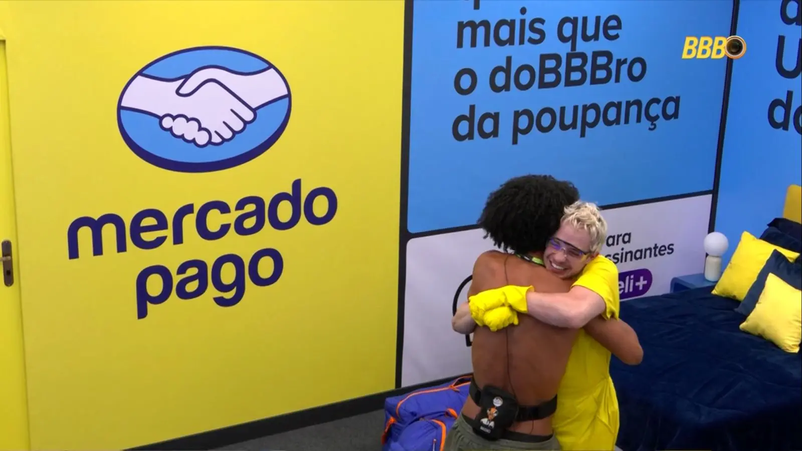 Ambiente colorido com paredes em tons de amarelo e azul, decorado com logotipos e textos promocionais. À esquerda, há um grande painel amarelo com um logotipo. À direita, há uma cama azul-marinho com travesseiros amarelos. No centro da cena, duas pessoas se abraçam; uma delas usa luvas amarelas e roupas da mesma cor, enquanto a outra está com as costas expostas e utiliza um equipamento preso à cintura.