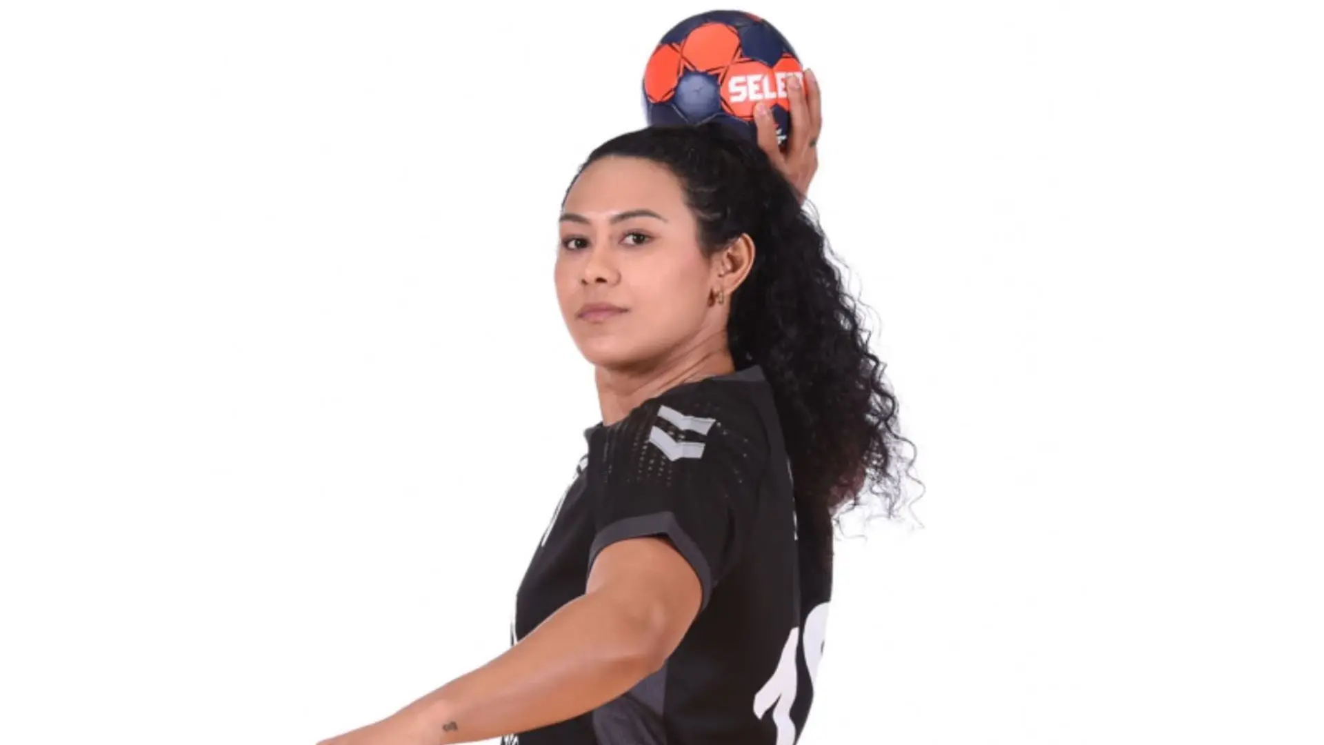Mulher jogadora de handebol segurando uma bola de handebol sobre a cabeça, vestindo uniforme preto, jogadora profissional de futebol feminino.