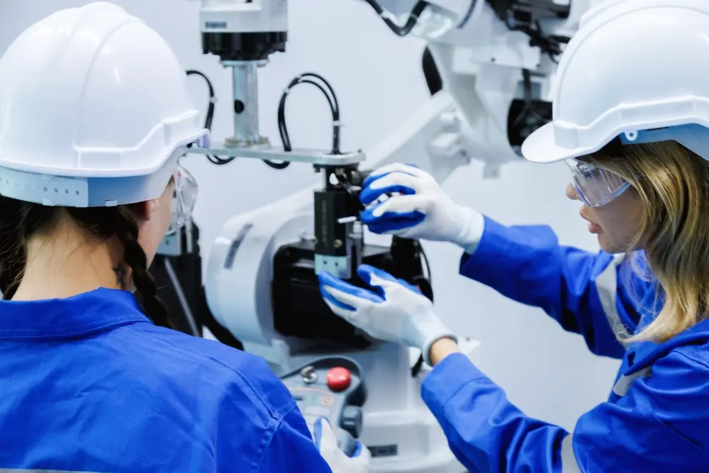 foto de duas mulheres jovens operando máquina. Elas usam equipamento de proteção na cabeça e luvas.