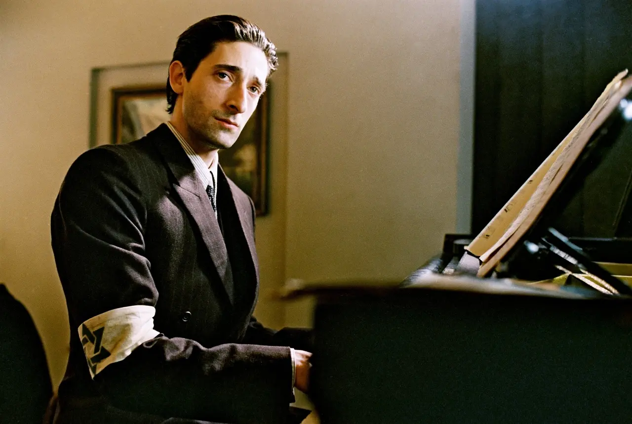O ator Adrien Brody em 'O Pianista' interpreta Wladyslaw Szpilman. Na imagem, sentado ao piano, ele veste um terno escuro com uma braçadeira da Estrela de Davi no braço direito enquanto olha seriamente para a câmera. A iluminação suave destaca seu perfil e as partituras sobre o instrumento, criando uma atmosfera melancólica e solene.