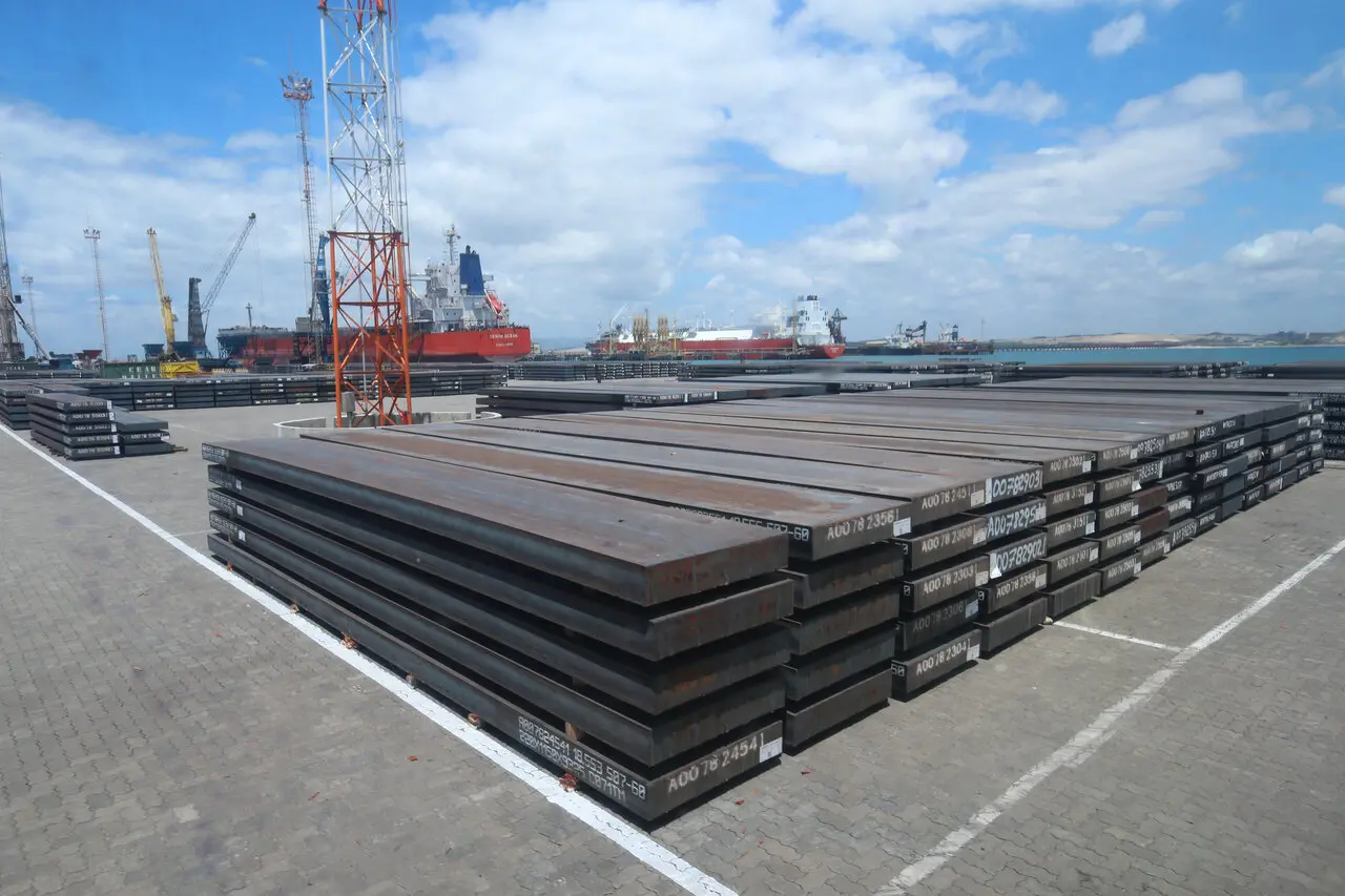 Pátio portuário com grandes chapas de aço empilhadas em primeiro plano. Ao fundo, há navios atracados, guindastes e estruturas industriais próximas ao mar, sob céu parcialmente nublado.