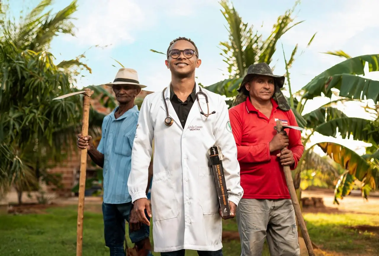 Na imagem, fotografia ao ar livre, em luz natural, mostrando três homens em um ambiente rural com vegetação tropical (bananeiras e palmeiras) ao fundo. Ao centro: Um homem negro jovem, sorridente, veste um jaleco branco de médico sobre uma camisa preta. Ele usa óculos de armação escura e carrega um estetoscópio no pescoço. Sob o braço esquerdo, segura um livro grande de capa preta intitulado 