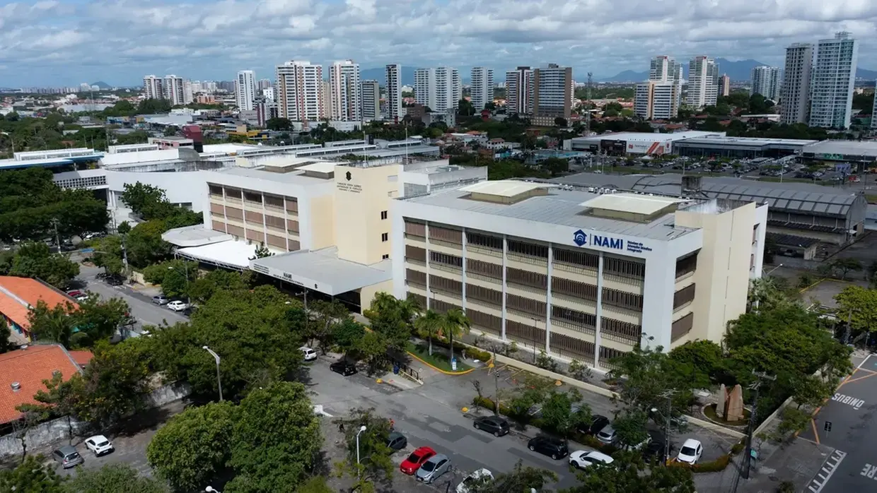 Na imagem, vista aérea do prédio do NAMI (Núcleo de Atenção Médica Integrada), na Universidade de Fortaleza (Unifor). O edifício possui arquitetura moderna em tons de branco e bege, com o logotipo azul do NAMI em destaque na lateral direita. À frente do prédio, há um estacionamento com diversos carros e áreas arborizadas. Ao fundo, observa-se o horizonte da cidade de Fortaleza com vários edifícios residenciais sob um céu levemente nublado.