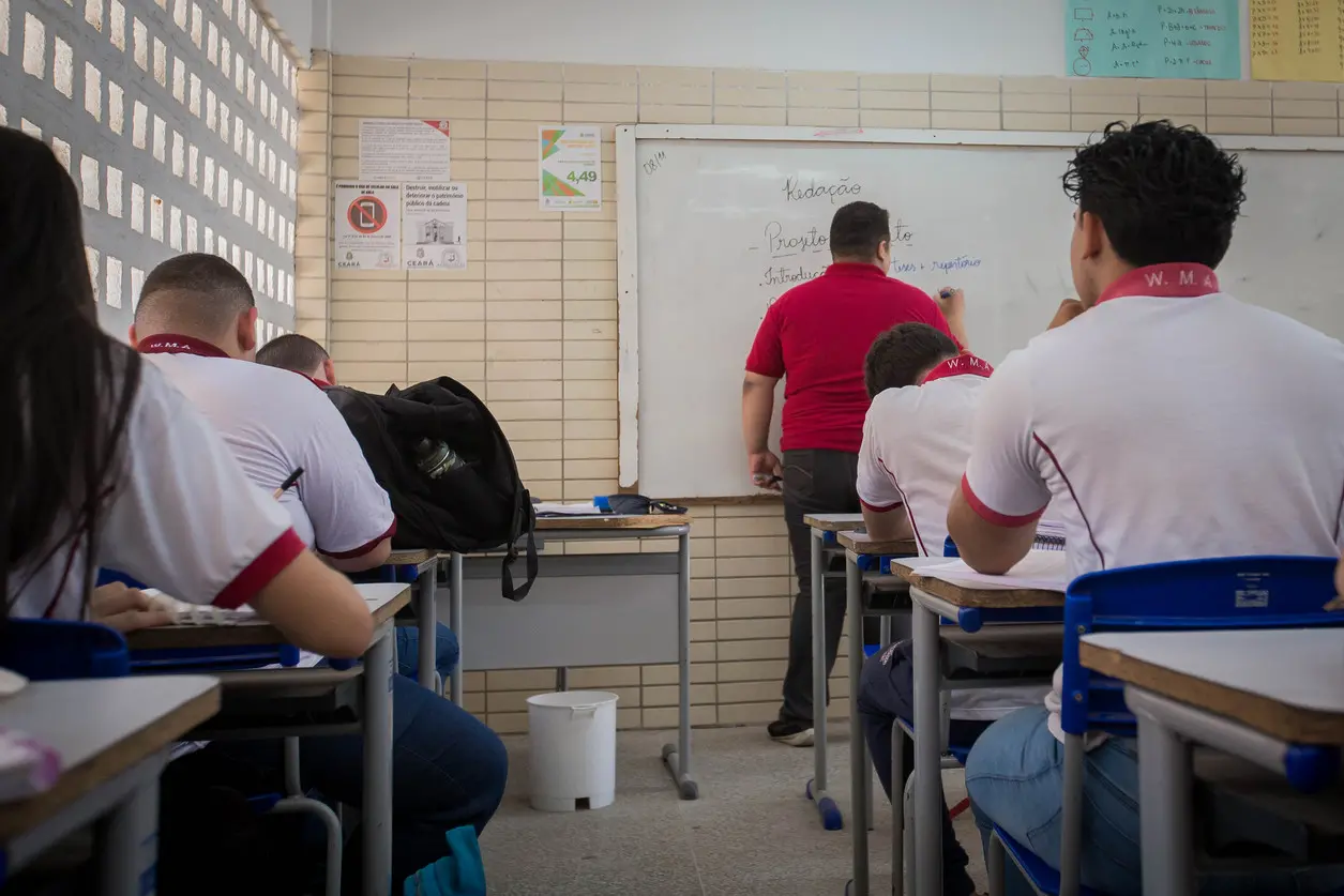 Professor escrevendo no quadro branco para alunos sentados em uma sala de aula.
