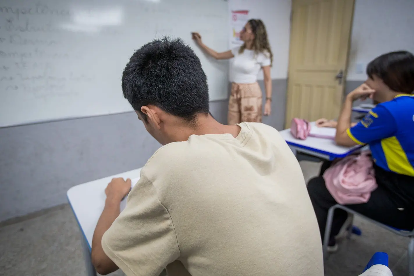 Vista de trás de um aluno em uma sala de aula, enquanto uma professora escreve no quadro branco e outro aluno assiste.