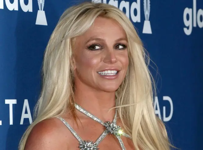 Britney é uma mulher branca e loira de cabelo liso, comprido. Na foto, ela está sorrindo, de vestido trançado no pescoço.