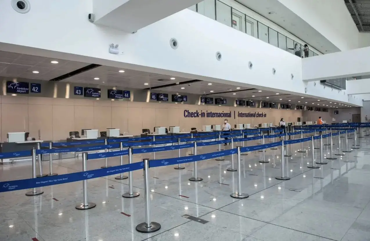 Área de check-in internacional de um aeroporto, com vários balcões alinhados e divisórias de fila azuis organizadas em um grande saguão amplo e bem iluminado. Há poucas pessoas ao fundo próximas aos balcões. Placas numeradas de 42 a 55 estão suspensas acima dos guichês.