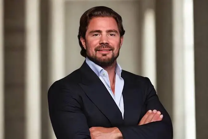 Daniel Vorcaro sorri de braços cruzados vestindo um paletó escuro sobre uma camisa azul clara desabotoada no colarinho. A imagem é de cunho profissional.