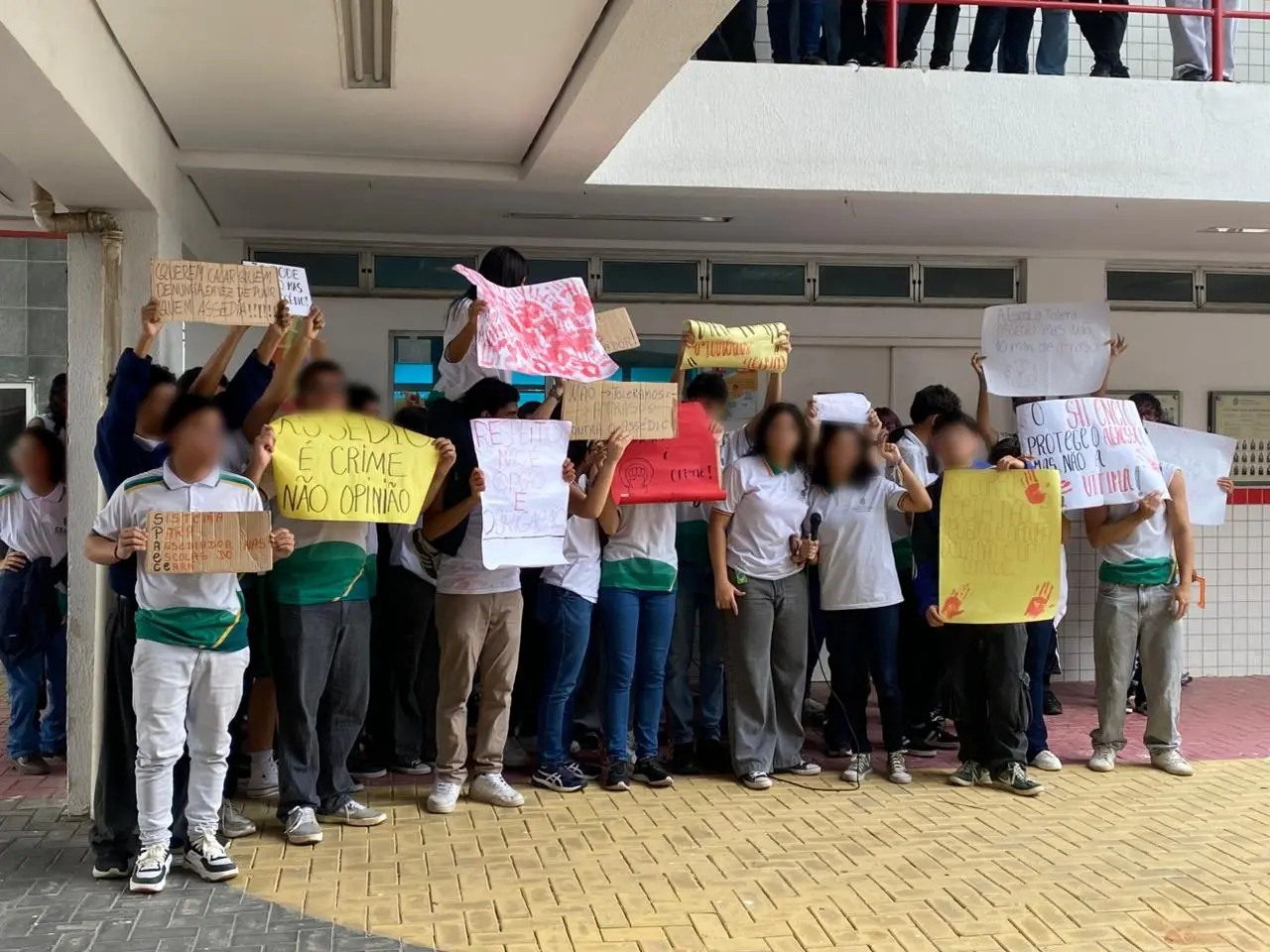 Um grupo de estudantes em um pátio escolar estadual no Ceará protestando contra o assédio e a violência, segurando cartazes com mensagens como 'Assédio é crime, não opinião' e 'O silêncio protege o agressor'.