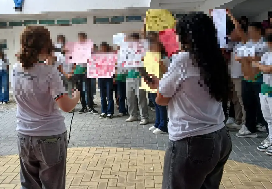 Duas meninas seguram cartazes e fazem falas com microfone durante protesto contra assédio sexual em uma escola. Elas estão cercadas por outros alunos fardados segurando cartazes com palavras de ordem.