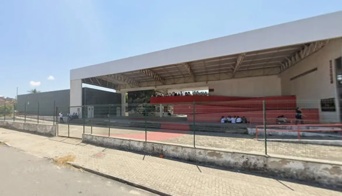 Fachada da escola Maria Angela, em Fortaleza, um bloco retangular branco com centro vazado e rampas vermelhas de acesso ao segundo andar. A unidade é cercada por grades e tem alunos sentados no chão do pátio.