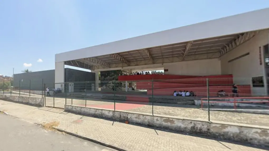 Fachada da escola Maria Angela, em Fortaleza, um bloco retangular branco com centro vazado e rampas vermelhas de acesso ao segundo andar. A unidade é cercada por grades e tem alunos sentados no chão do pátio.