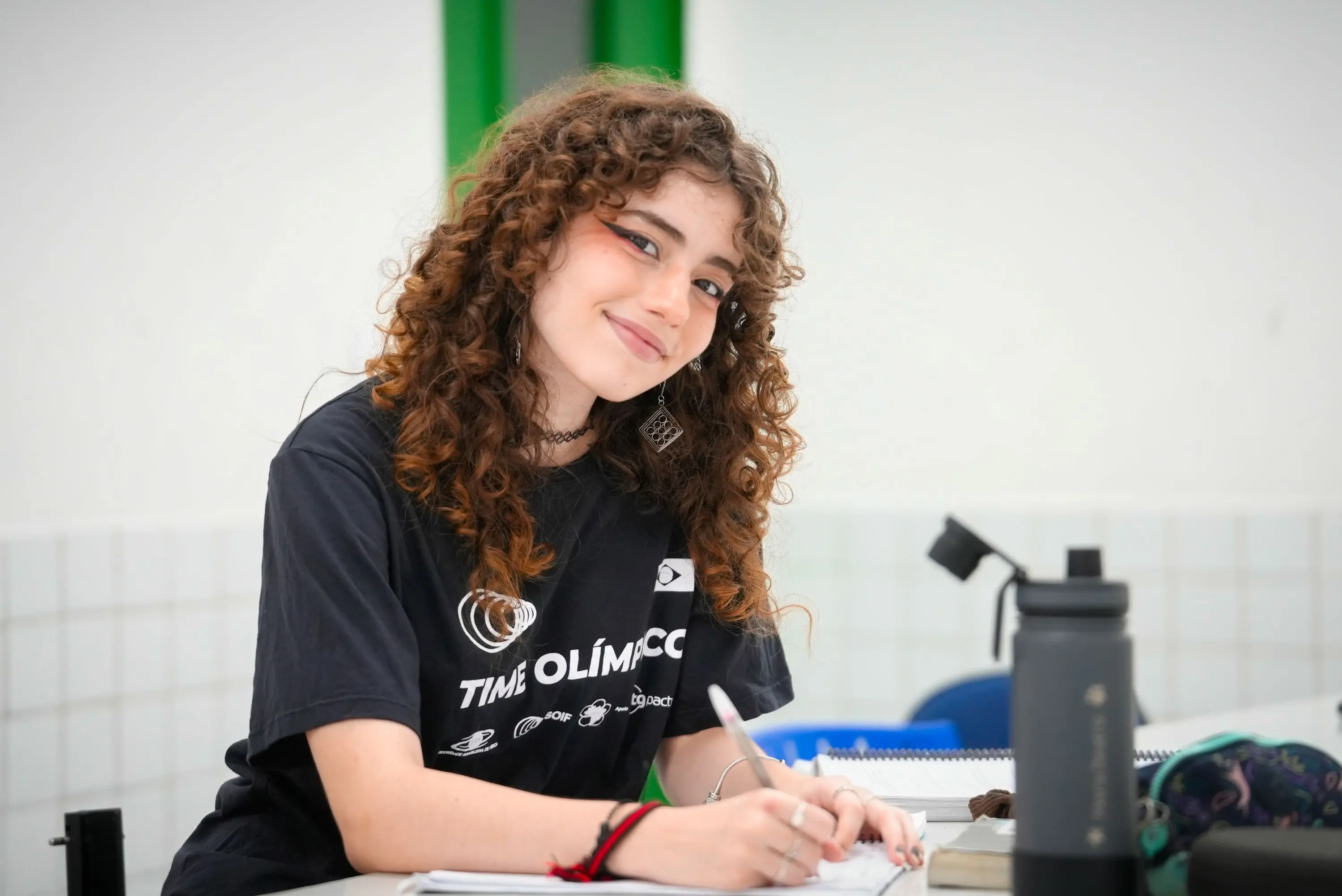 Jovem estudante com cabelos cacheados sorrindo enquanto escreve em um caderno, vestindo uma camiseta TIME OLÍMPICO em sala de aula.
