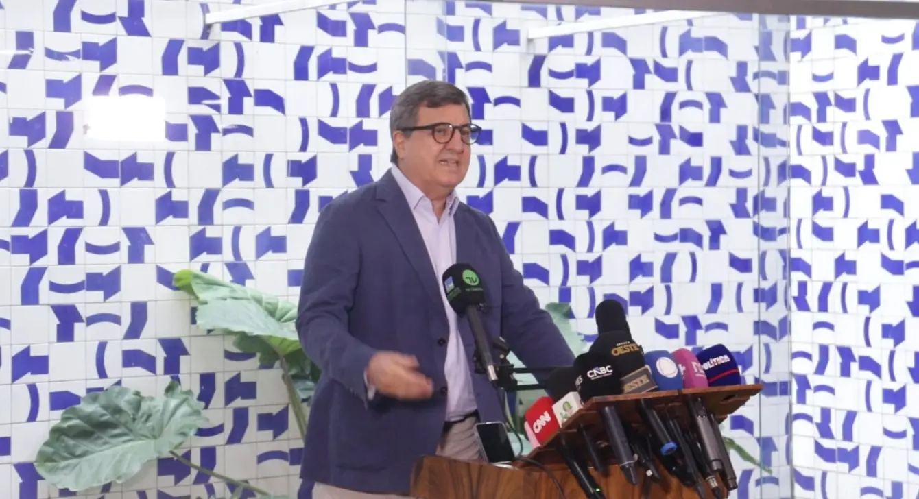 Danilo Forte usa óculos e terno azul fala em um púlpito durante uma coletiva de imprensa, cercado por vários microfones de diferentes emissoras, com uma parede de azulejos brancos e azuis ao fundo e plantas ao lado.