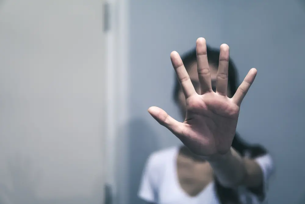 Na imagem, fotografia em tons frios de uma mulher com o rosto desfocado ao fundo, estendendo a palma da mão aberta em direção à câmera em um gesto de 'pare' ou sinal de interrupção.
