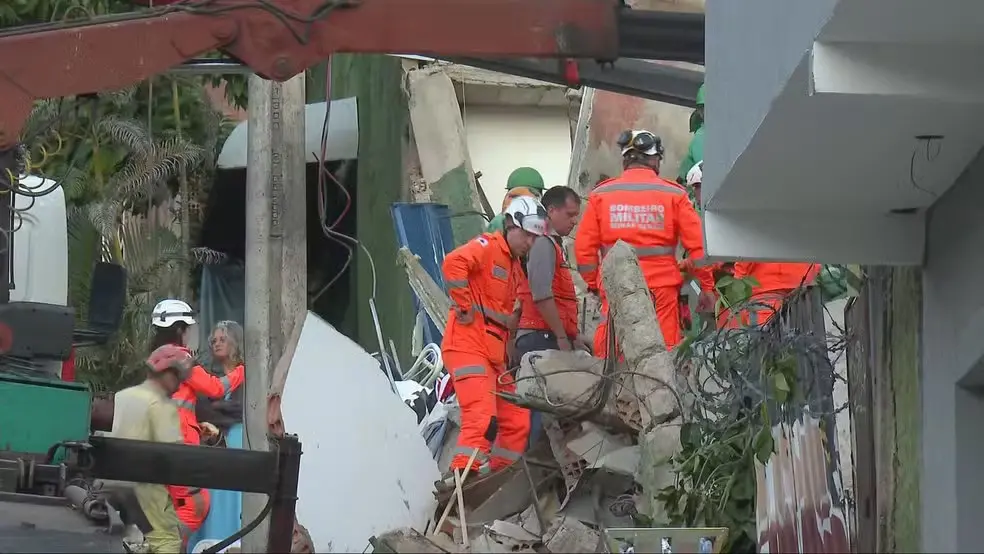 Equipes de resgate em ação após desabamento de estrutura, com profissionais usando equipamentos de segurança e colocando placa de contenção em uma área de risco.