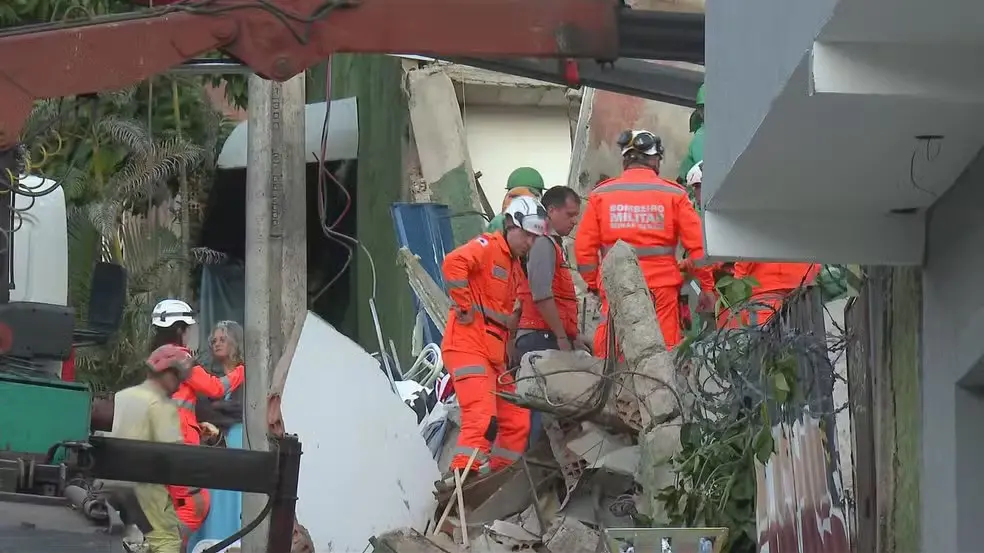 Equipes de resgate em ação após desabamento de estrutura, com profissionais usando equipamentos de segurança e colocando placa de contenção em uma área de risco.