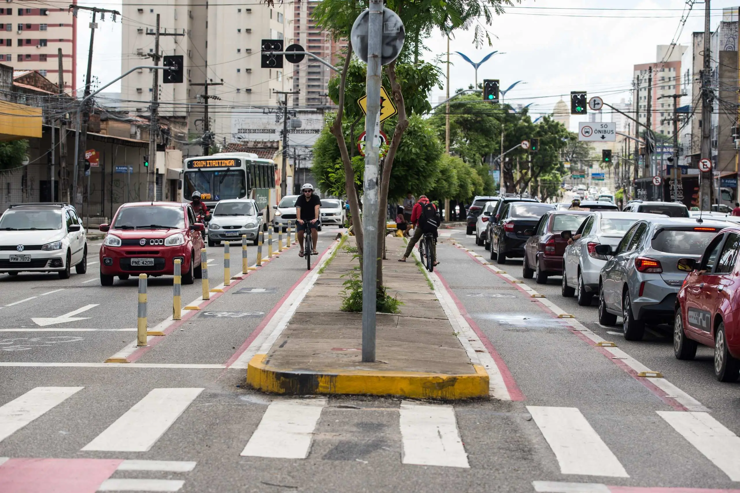 Esta fotografia captura uma avenida movimentada em um dia nublado, com foco em uma ciclofaixa central de sentido duplo. A via para ciclistas é delimitada por linhas vermelhas no asfalto e separada dos carros por pequenos balizadores amarelos e brancos; nela, dois ciclistas pedalam em direções opostas. À esquerda, o tráfego flui com carros brancos e vermelhos e um ônibus urbano branco ao fundo. À direita, há uma fila de veículos estacionados ou em trânsito lento. No centro, um canteiro divisor estreito e elevado possui árvores jovens e postes de sinalização. Em primeiro plano, as listras brancas de uma faixa de pedestres cruzam a base da imagem, enquanto ao fundo observam-se prédios residenciais, semáforos e uma rede de fios elétricos que atravessa o céu cinzento.