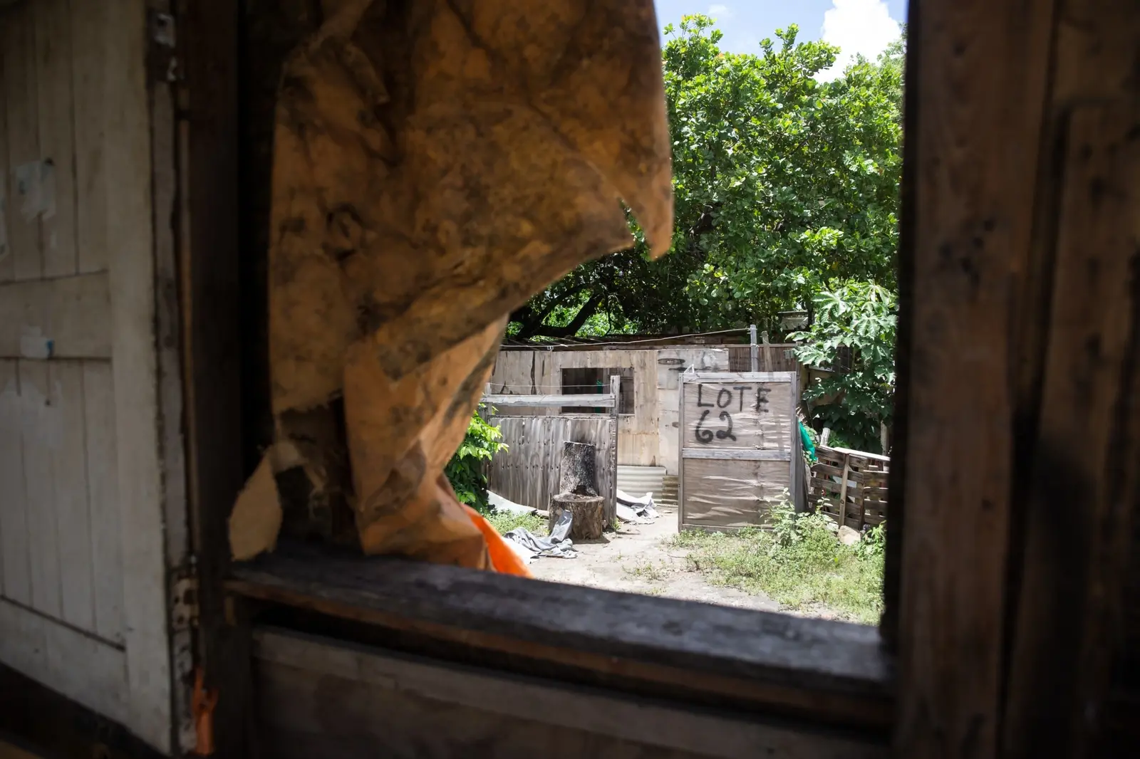 Visão através de uma janela quebrada mostrando um lote demarcado com LOTE 62 escrito em uma placa de madeira.