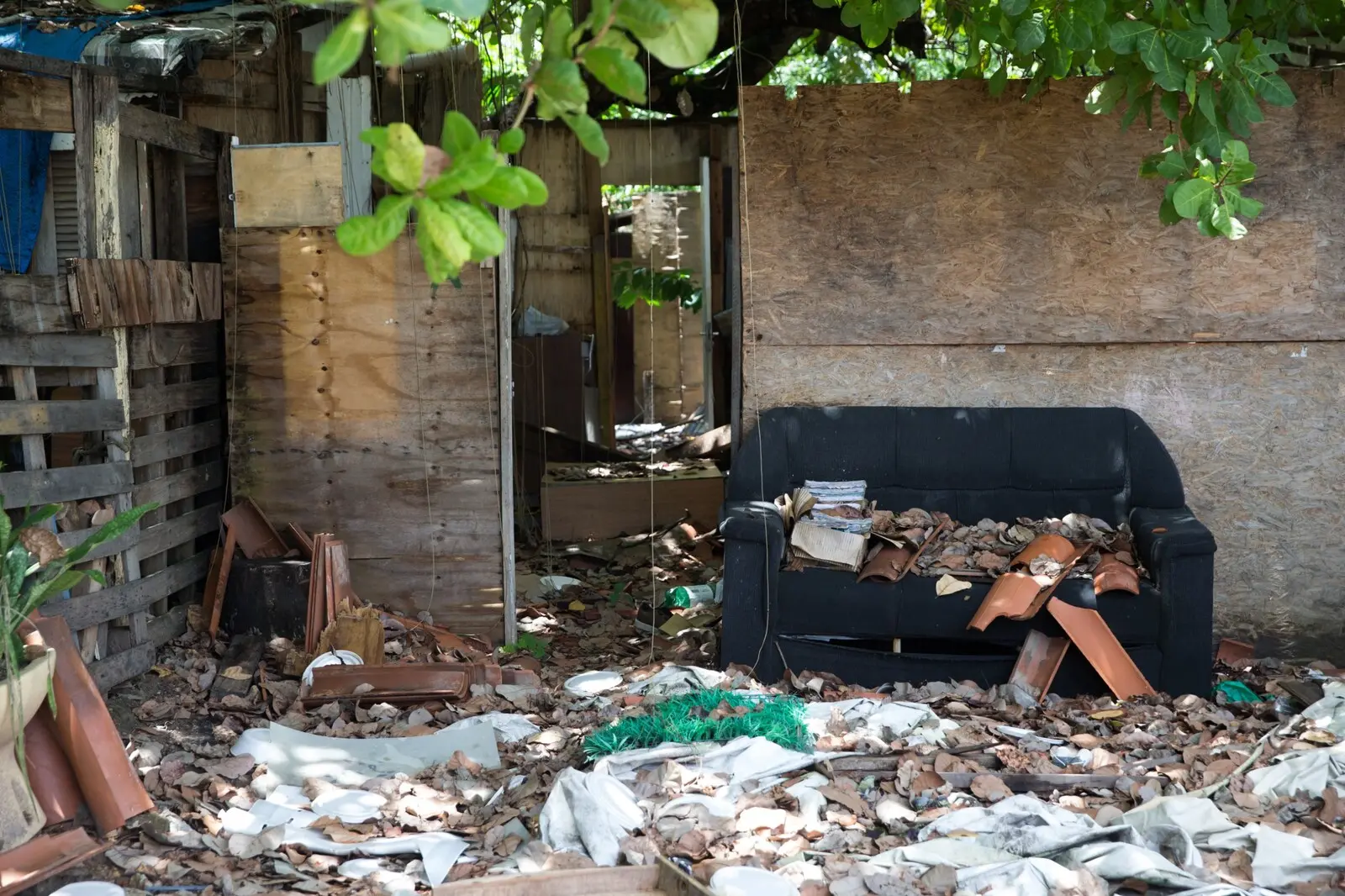 Interior de um abrigo destruído com um sofá preto coberto de telhas quebradas, folhas secas e lixo espalhado pelo chão.