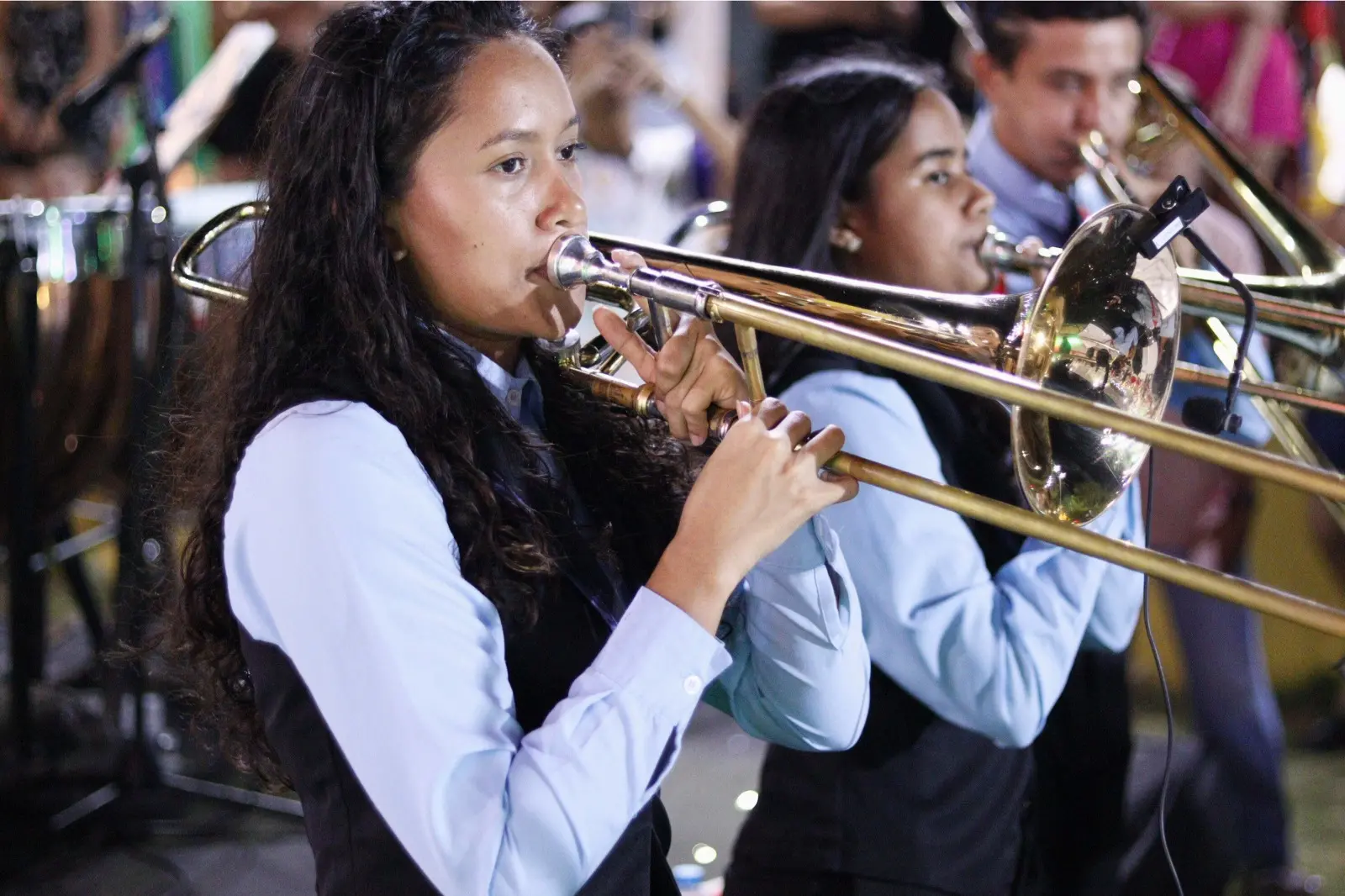 Imagem mostra Ana Cristina em destaque tocando um instrumento de sopro dourado usando uma camisa social de manga longa azul claro com outras pessoas tocando instrumentos ao fundo.