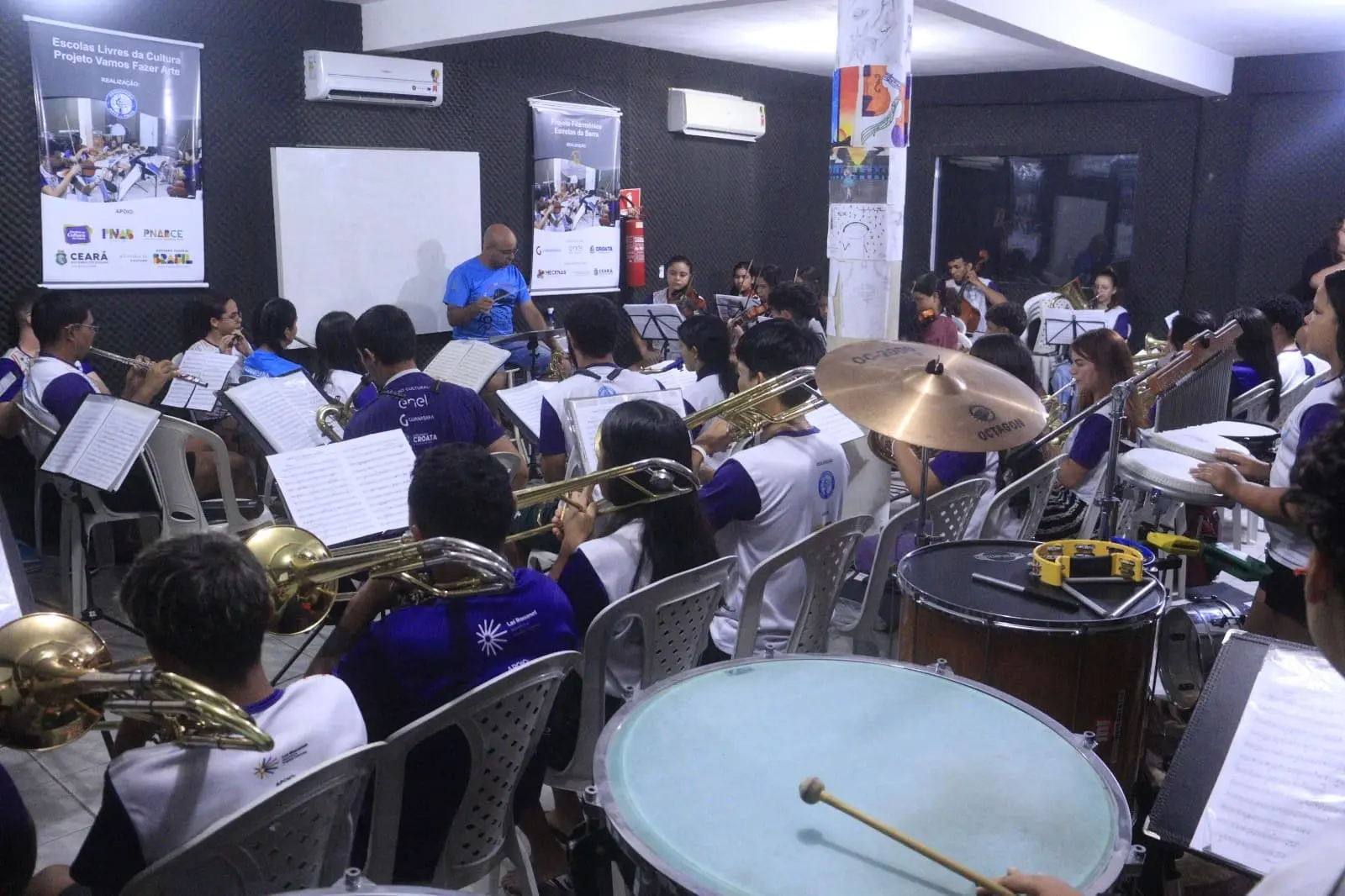 Imagem mostra jovens tocando diversos instrumentos em uma orquestra ensaiando dentro de uma sala com um maestro à frente.