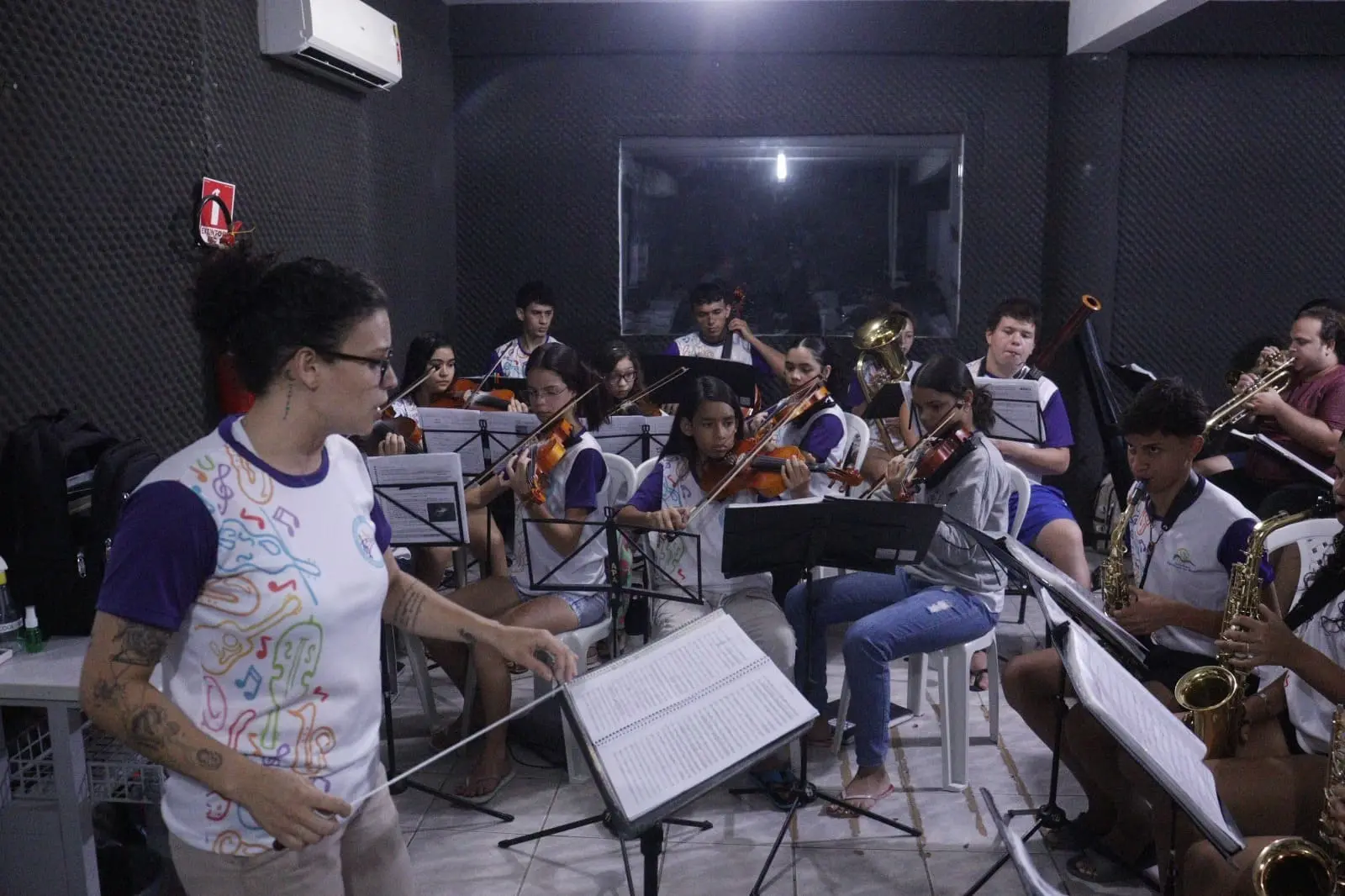 Imagem mostra jovens tocando diversos instrumentos em uma orquestra ensaiando dentro de uma sala com um maestro à frente.