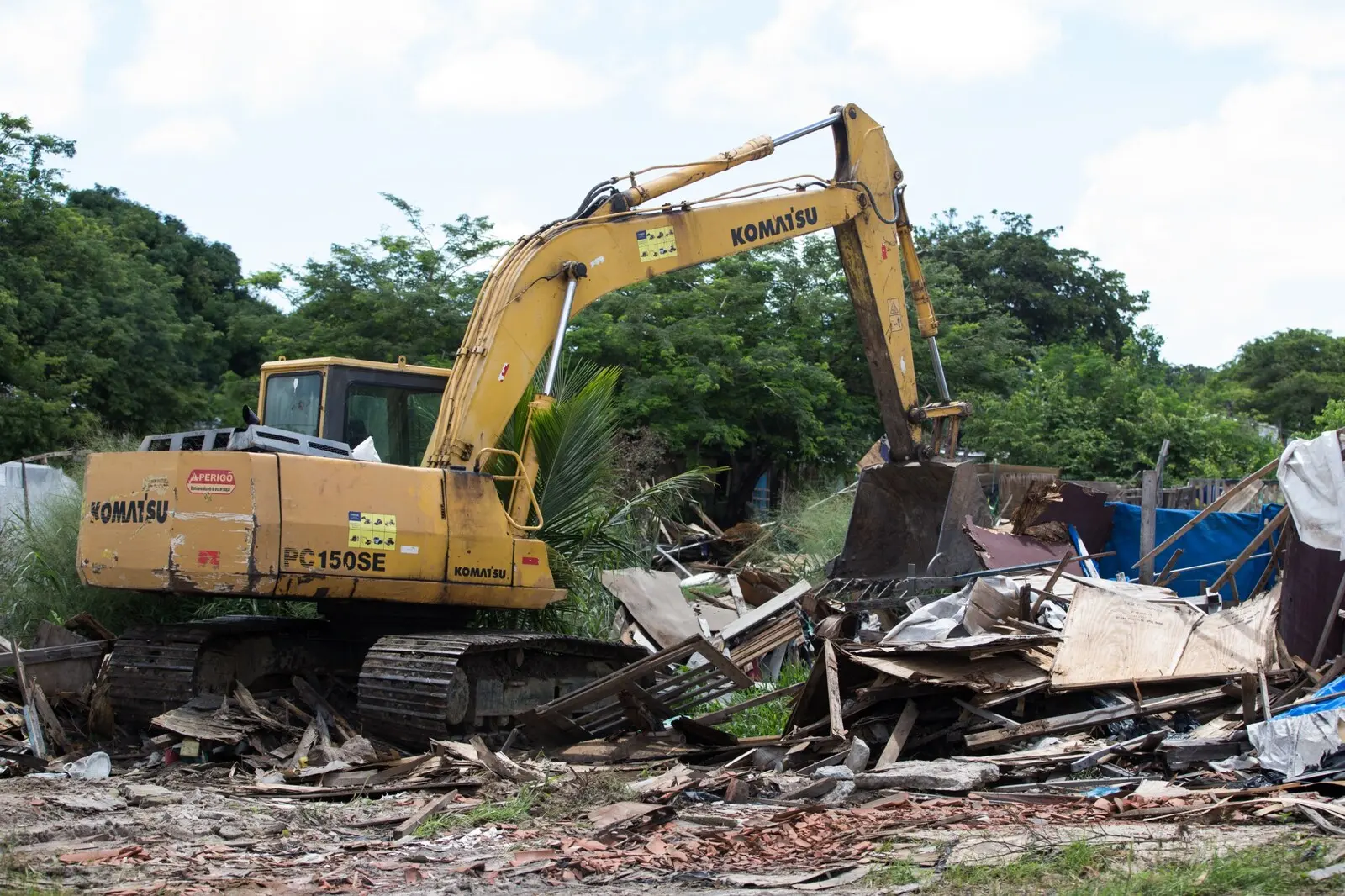 Escavadeira amarela da marca Komatsu demolindo barracos de madeira e entulho em um terreno com vegetação ao fundo em Fortaleza.
