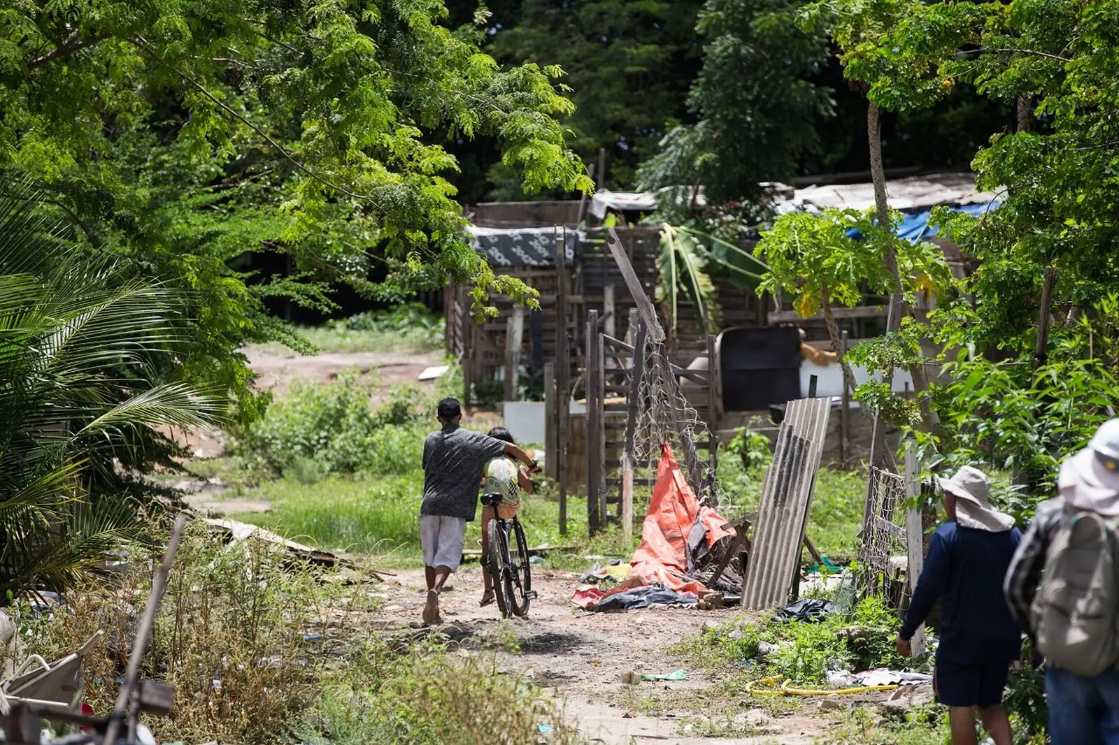Homem e criança em uma bicicleta percorrem caminho de terra em ocupação precária, cercada por vegetação e barracos improvisados.
