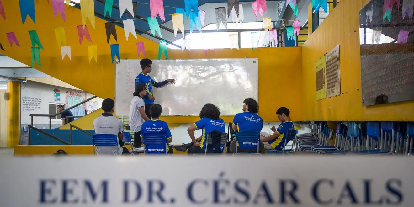 Professor em pé diante de um quadro branco dá explicação para um grupo de estudantes sentados em cadeiras dentro de um espaço escolar colorido, decorado com bandeirinhas no teto. A cena ocorre em área aberta da escola, com paredes amarelas e outros alunos ao fundo.