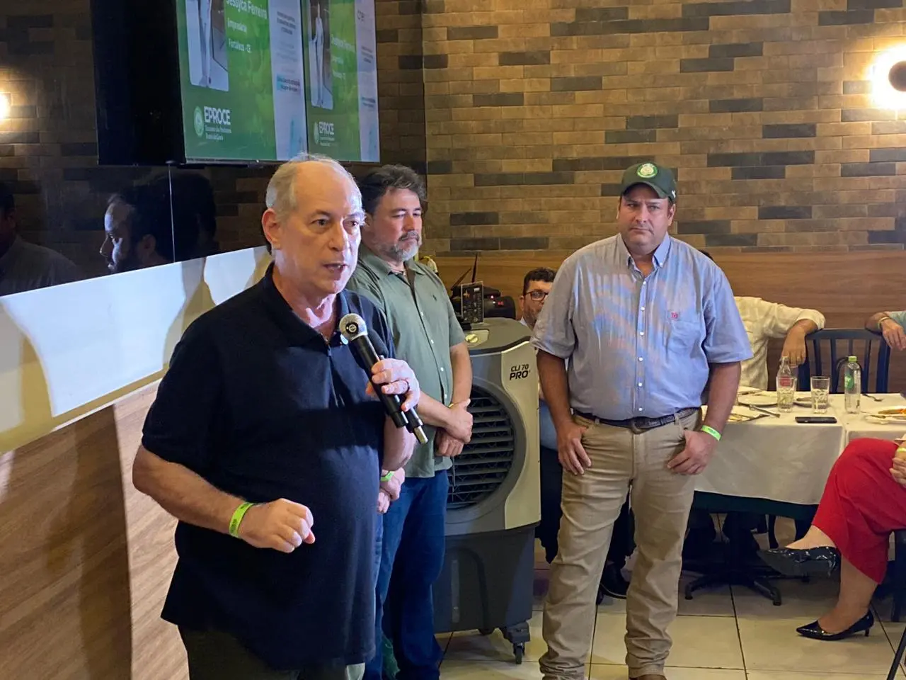 Homem fala ao microfone durante evento em ambiente interno com paredes de madeira. Trata-se de Ciro Gomes. Ao fundo, dois homens estão em pé observando, enquanto outras pessoas estão sentadas em mesas. Uma tela exibe uma apresentação ao fundo e há um climatizador portátil no local.