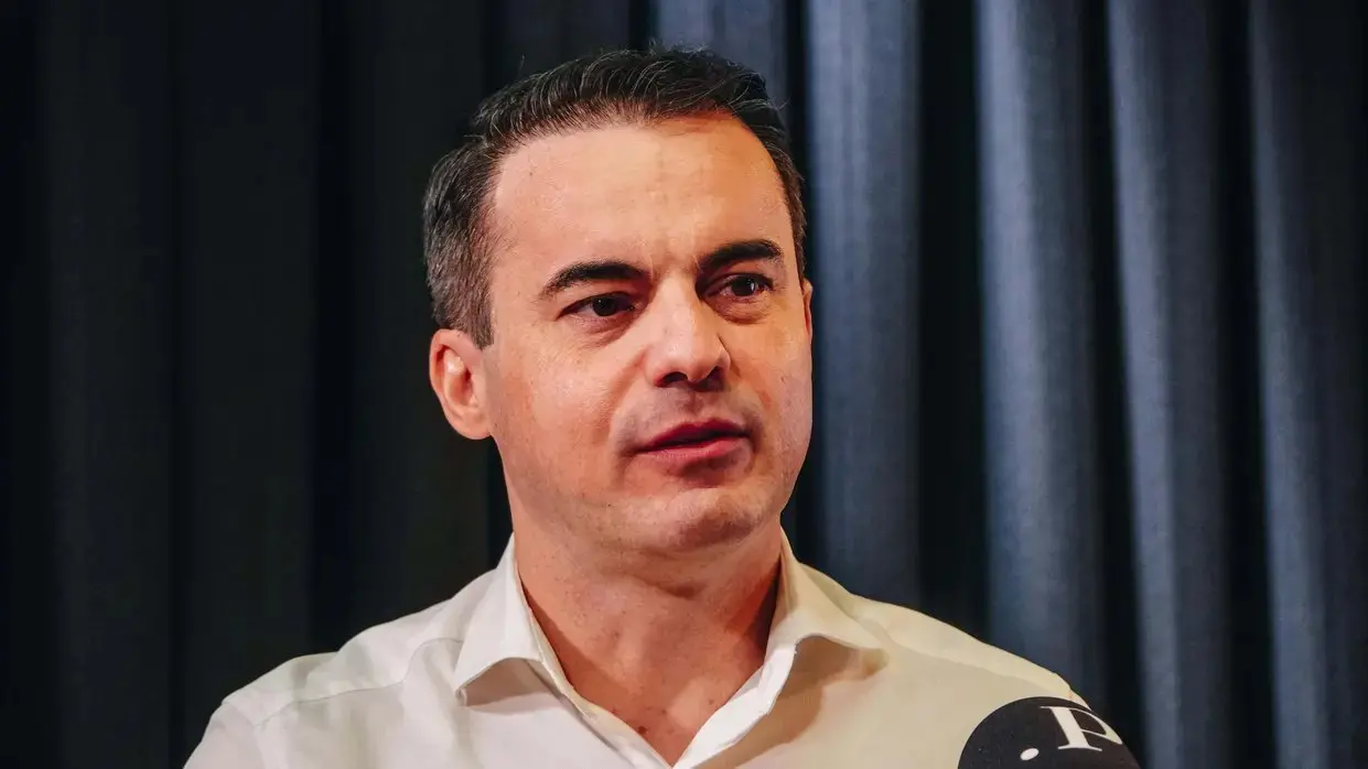 Homem de meia-idade com cabelo curto escuro e grisalho nas laterais, usando camisa branca, olhando levemente para o lado durante uma entrevista, com um microfone visível na parte inferior da imagem e fundo de cortinas escuras.