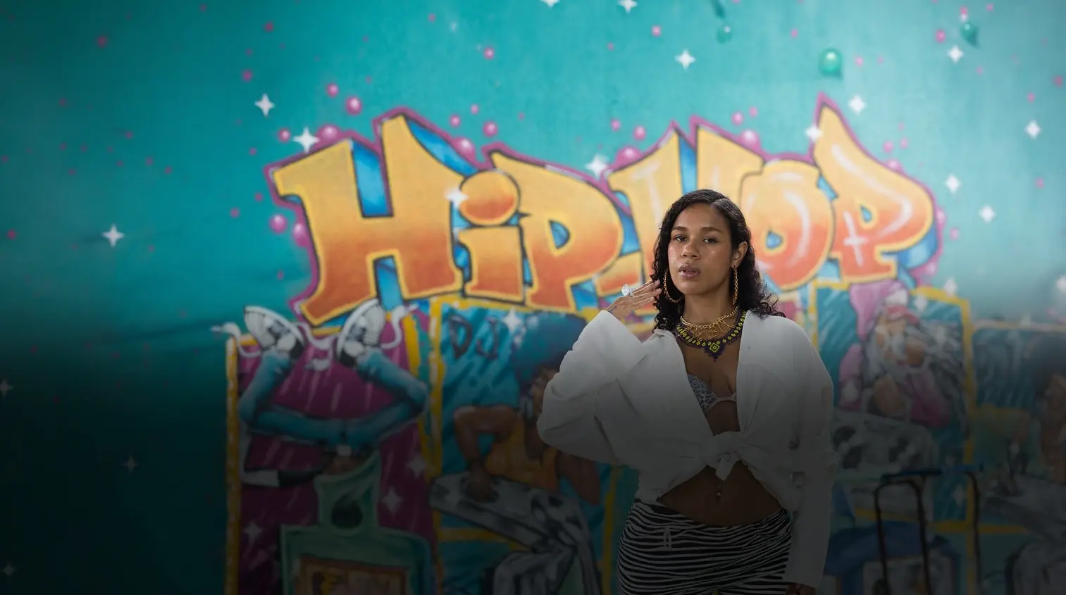 Foto da rapper Cabulosa em frente a um grafite onde se lê 