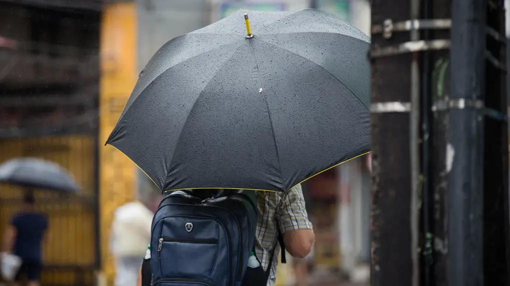 Um pedestre caminha sob chuva usando um guarda-chuva preto molhado e carregando uma mochila azul nas costas. Ao fundo, o cenário urbano aparece desfocado com tons de amarelo e cinza.