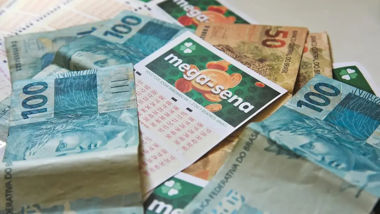 Um volante da Mega-Sena está posicionado centralmente sobre diversas cédulas de Real de 50 e 100 espalhadas. A imagem destaca o contraste entre o papel da loteria e o dinheiro em espécie em uma composição de plano próximo.