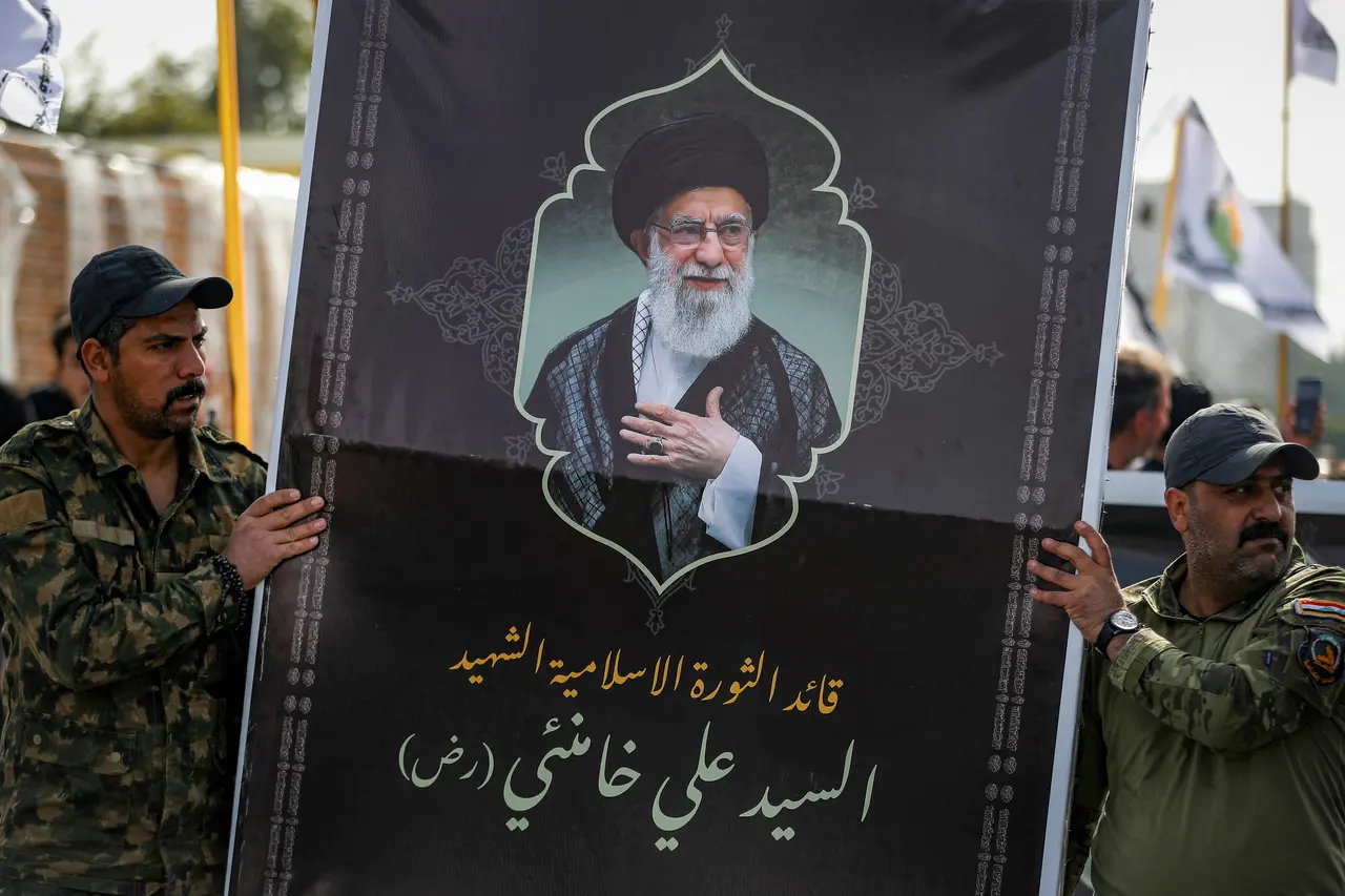 Dois homens vestindo uniformes militares camuflados seguram um grande cartaz preto com a imagem do aiatolá Ali Khamenei. O cartaz exibe o líder religioso ao centro e inscrições em árabe na parte inferior.