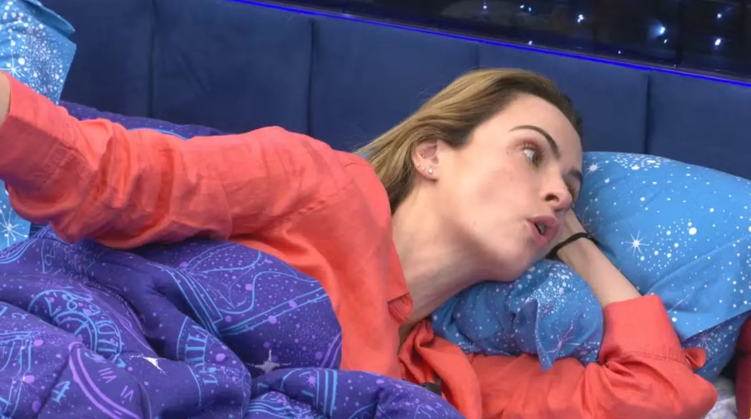 Mulher deitada na cama, apoiada em um travesseiro azul com estampa de estrelas, usando uma camisa laranja e olhando para o lado enquanto fala. Ela está coberta por um edredom roxo com estampas astronômicas, em um quarto com cabeceira azul e iluminação suave ao fundo.