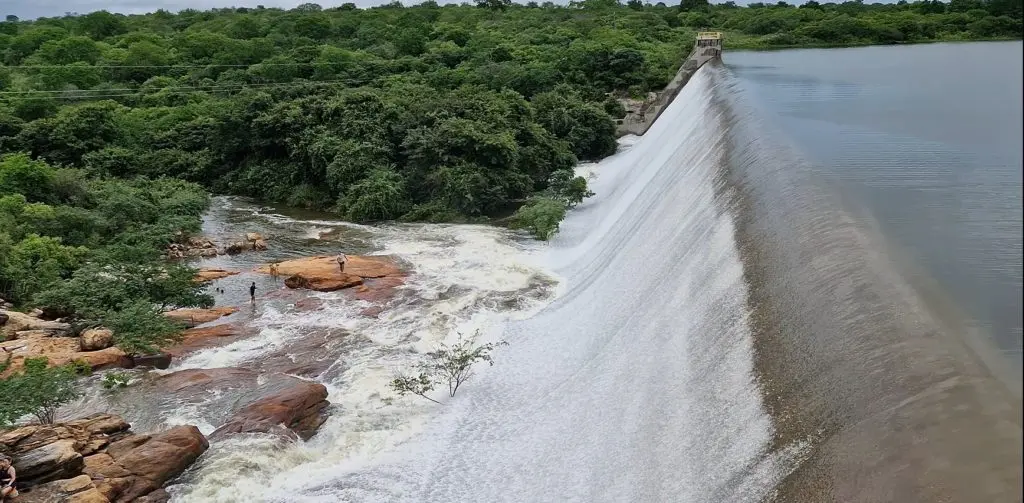 Foto aérea de um açude sangrando muita água por cima da parede, inundando um vale rochoso abaixo e rodeado por áreas verdes.