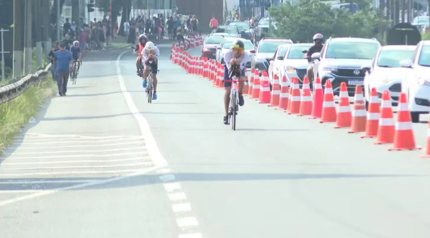 Ciclistas participam de uma prova em via urbana delimitada por cones laranjas, enquanto carros seguem na faixa ao lado e pedestres observam ao fundo.