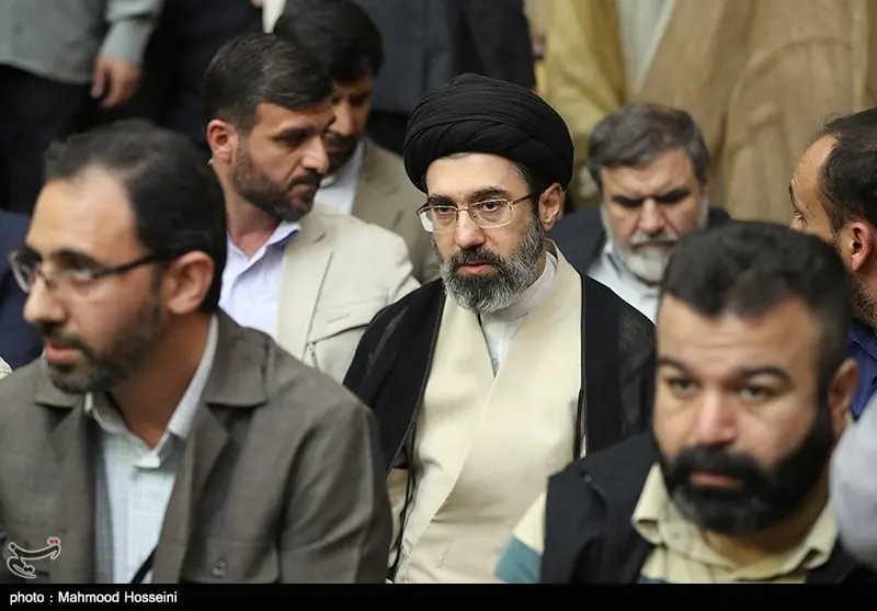 Reunião de homens islâmicos com destaque central para Mojtaba Khamenei, um homem de 50 anos com barba grisalha usando óculos e turbante preto.
