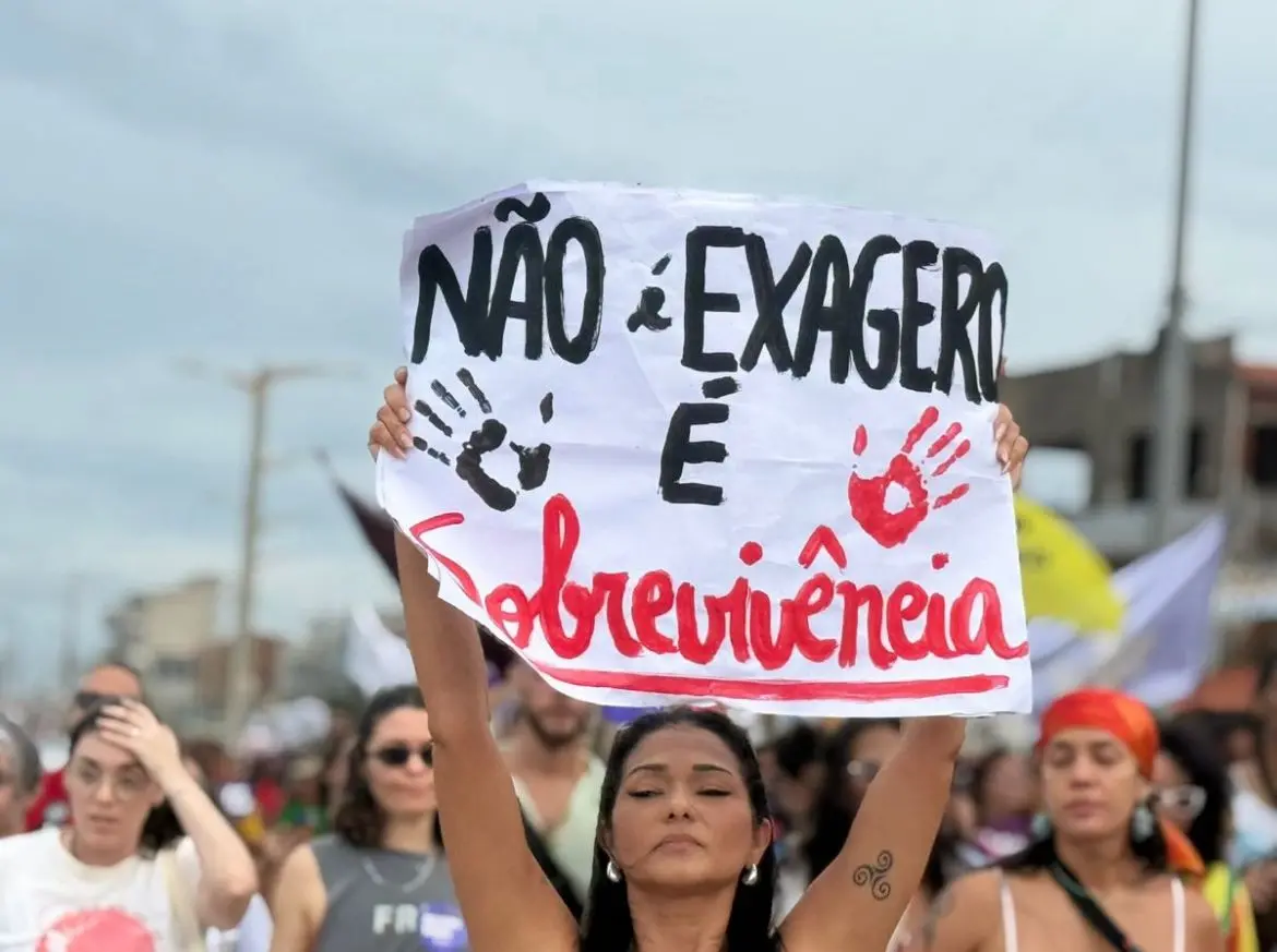 Mulher segura acima da cabeça um cartaz escrito “Não é exagero, é sobrevivência”, com marcas de mãos pintadas em preto e vermelho. Ela participa de uma manifestação ao lado de outras mulheres em uma rua, durante o dia. Ao fundo, aparecem pessoas caminhando juntas no protesto e parte da paisagem urbana desfocada.