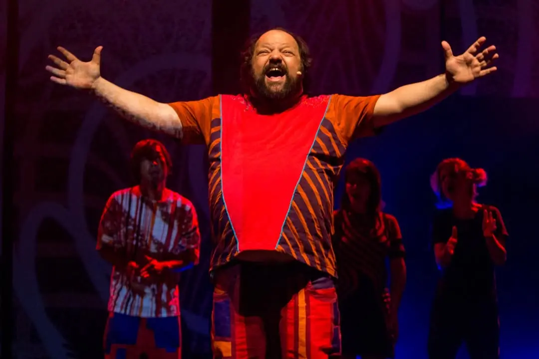 Cena de um espetáculo teatral com ator em destaque usando roupa colorida, na frente de um grupo de pessoas, sob iluminação de palco.